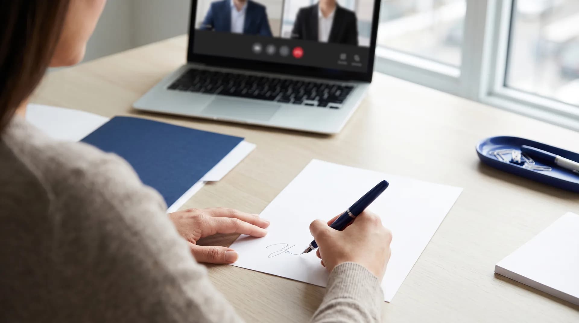 A person signs a document at a bright desk while a commissioner appears on a video call on their laptop